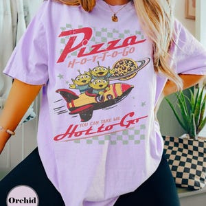 May include: A lilac t-shirt with a retro graphic. The design includes the text "Pizza H-O-T-T-O-Go", a rocket ship, three green aliens, and a pizza planet. The phrase "You can take me Hot to Go" is also featured.