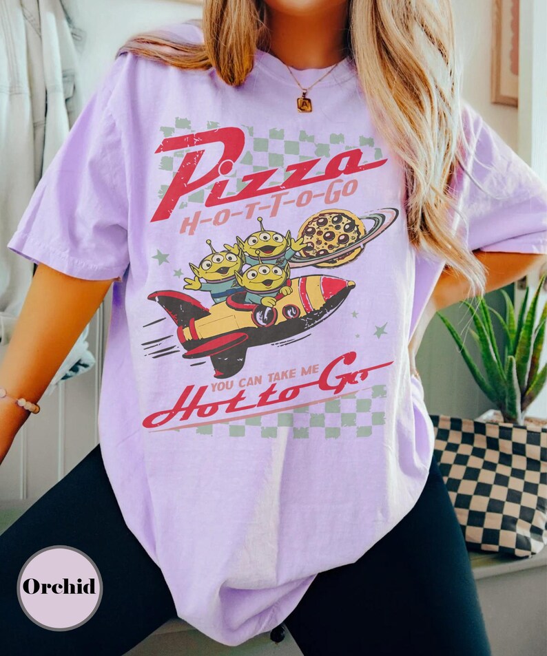 May include: A lilac t-shirt with a retro graphic. The design includes the text "Pizza H-O-T-T-O-Go", a rocket ship, three green aliens, and a pizza planet. The phrase "You can take me Hot to Go" is also featured.