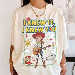 May include: A cream-colored t-shirt featuring a graphic of the character Jessie from Toy Story playing an acoustic guitar. The text reads I KNEW IT, I KNEW YOU. The design includes a cowboy hat, boots, a star badge, clouds, and colorful blocks.