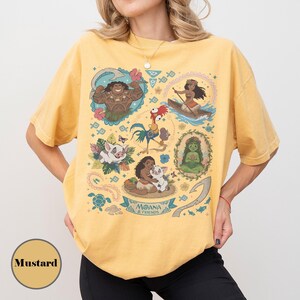May include: Mustard-colored t-shirt featuring a colorful graphic of characters from the animated film Moana. The design includes illustrations of Maui, Moana, Pua, Hei Hei, and other elements from the movie. The words "Moana & Friends" are also visible.