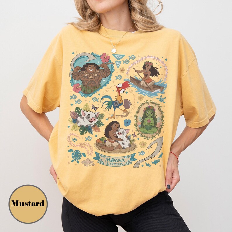 May include: Mustard-colored t-shirt featuring a colorful graphic of characters from the animated film Moana. The design includes illustrations of Maui, Moana, Pua, Hei Hei, and other elements from the movie. The words "Moana & Friends" are also visible.