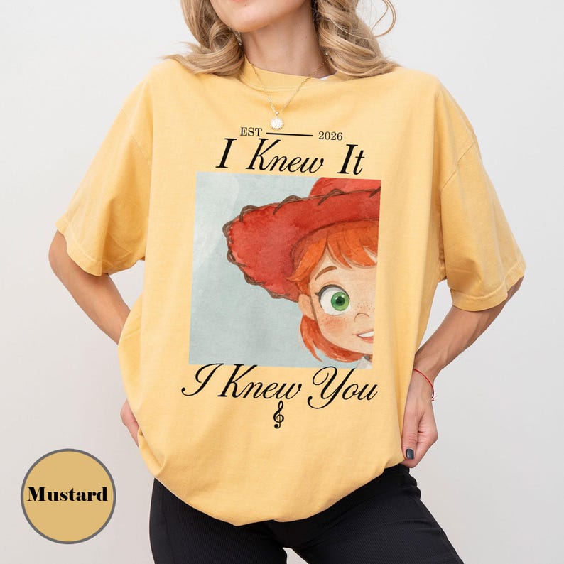 May include: A mustard yellow short-sleeve t-shirt featuring a graphic print of a red-hatted character with green eyes. The text reads "EST 2026 I Knew It I Knew You" with a treble clef symbol. The shirt has a crew neckline and a relaxed fit.