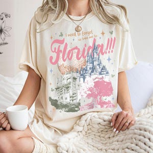 May include: Cream-colored t-shirt with a graphic design featuring the word "Florida!!!" in pink, along with illustrations of a castle, a sphere, and a tree. The shirt also has the text "I need to forget so take me to..." in a smaller font.