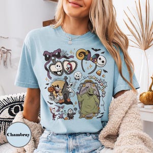 Op de afbeelding: Lichtblauw T-shirt met een ontwerp met personages uit "The Nightmare Before Christmas". Het ontwerp bevat Jack Skellington, Sally, Oogie Boogie en andere iconische beelden. Het woord "Chambray" staat linksonder gedrukt.
