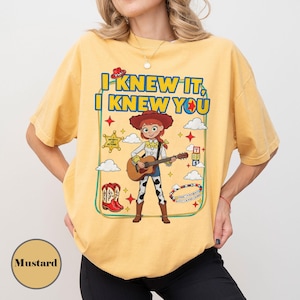 May include: A mustard yellow t-shirt featuring a graphic of a cowgirl character playing an acoustic guitar. The text reads I KNEW IT, I KNEW YOU. The design includes a sheriff star, red cowboy boots, clouds, stars, and a beaded bracelet graphic.