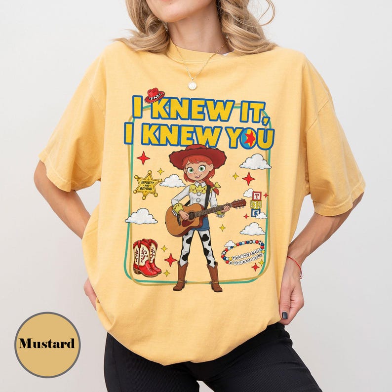 May include: A mustard yellow t-shirt featuring a graphic of a cowgirl character playing an acoustic guitar. The text reads I KNEW IT, I KNEW YOU. The design includes a sheriff star, red cowboy boots, clouds, stars, and a beaded bracelet graphic.