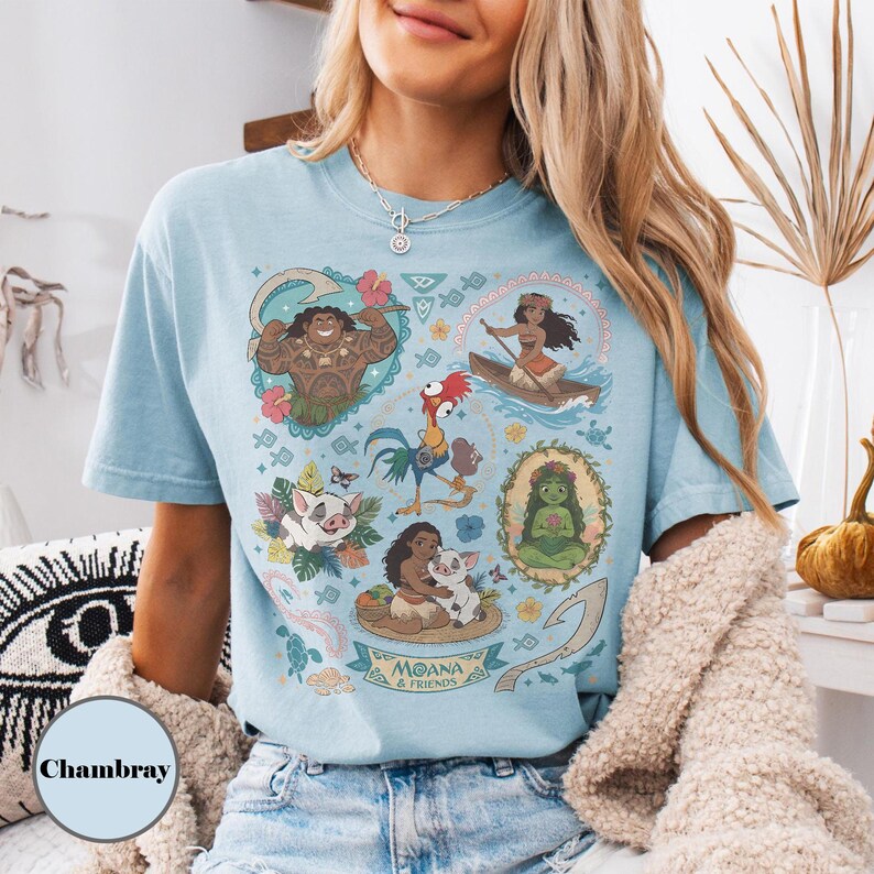 May include: Light blue t-shirt featuring a colorful graphic of characters from the animated film "Moana." The design includes illustrations of Maui, Moana, Pua, Hei Hei, and other characters. The shirt has a crew neck and short sleeves.
