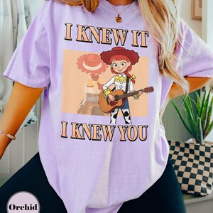 May include: A light purple short-sleeve t-shirt featuring a graphic of a cartoon cowgirl with red hair playing an acoustic guitar. The text "I KNEW IT" appears above the character and "I KNEW YOU" appears below, both in a bold, orange-outlined font.