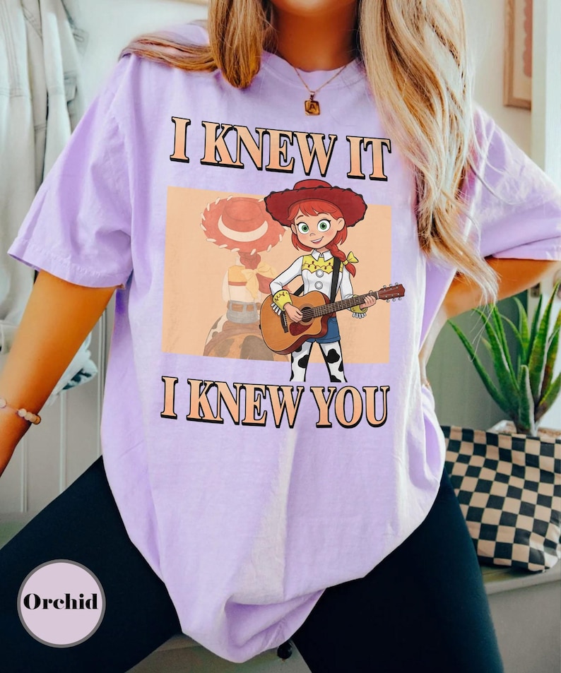May include: A light purple short-sleeve t-shirt featuring a graphic of a cartoon cowgirl with red hair playing an acoustic guitar. The text "I KNEW IT" appears above the character and "I KNEW YOU" appears below, both in a bold, orange-outlined font.