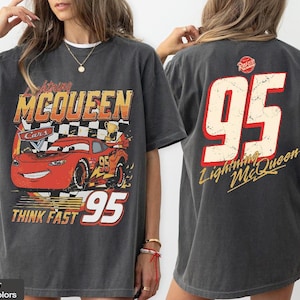 May include: Dark grey t-shirt featuring a graphic of Lightning McQueen from the Cars movie. The front design includes the text "Lightning McQueen Cars 95 Think Fast". The back of the shirt displays the number "95" and "Lightning McQueen".