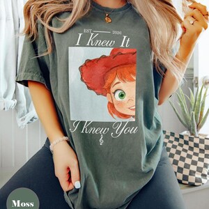 May include: A moss green short-sleeve t-shirt featuring a graphic of a cartoon character with red hair and a red cowboy hat. The text reads EST 2026, I Knew It, and I Knew You with a treble clef symbol below the text. The design is centered on the chest.