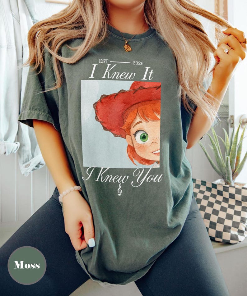 May include: A moss green short-sleeve t-shirt featuring a graphic of a cartoon character with red hair and a red cowboy hat. The text reads EST 2026, I Knew It, and I Knew You with a treble clef symbol below the text. The design is centered on the chest.