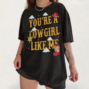 Puede incluir: Una camiseta negra de manga corta con el texto amarillo "YOU'RE A COWGIRL LIKE ME" y gr&aacute;ficos de un sombrero de vaquero rojo, una estrella de sheriff dorada, botas vaqueras rojas y nubes blancas en la parte frontal de la prenda.