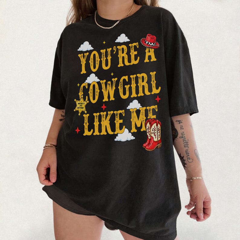 Puede incluir: Una camiseta negra de manga corta con el texto amarillo "YOU'RE A COWGIRL LIKE ME" y gr&aacute;ficos de un sombrero de vaquero rojo, una estrella de sheriff dorada, botas vaqueras rojas y nubes blancas en la parte frontal de la prenda.