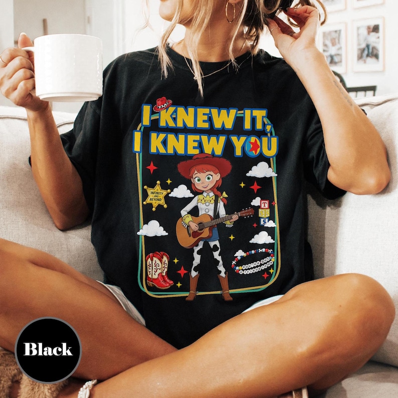 May include: A black t-shirt featuring a graphic print of a cowgirl character playing an acoustic guitar. The text reads I KNEW IT, I KNEW YOU. The design includes a sheriff star, red cowboy boots, colorful blocks, and a beaded bracelet graphic.
