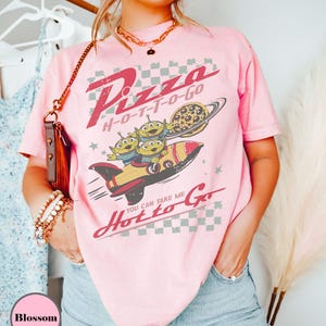 May include: Pale pink t-shirt with a retro graphic design. The design features the words "Pizza H-O-T-T-O-Go", a rocket ship with three green aliens, and a pizza-shaped planet. The shirt has short sleeves and a relaxed fit.