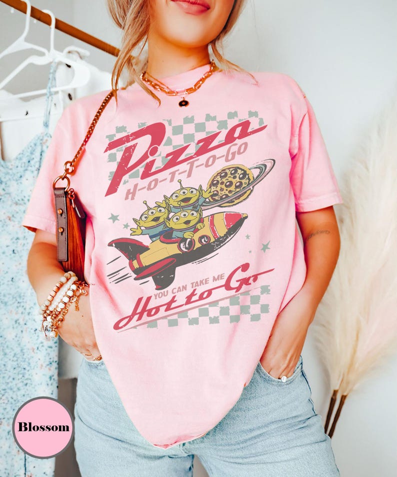 May include: Pale pink t-shirt with a retro graphic design. The design features the words "Pizza H-O-T-T-O-Go", a rocket ship with three green aliens, and a pizza-shaped planet. The shirt has short sleeves and a relaxed fit.