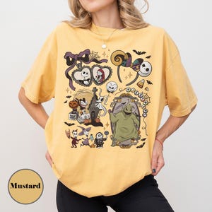Op de afbeelding: Mosterdkleurig T-shirt met een kleurrijke afbeelding van personages en elementen uit een populaire animatiefilm. Het ontwerp bevat Jack Skellington, Sally, Zero en Oogie Boogie, samen met vleermuizen, snoepma&iuml;s en andere spookachtige elementen.