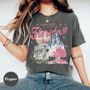 May include: Dark gray t-shirt with a colorful graphic of Florida landmarks, including a sphere, castle, and tree. The text "I need to forget so take me to... Florida!!!" is printed in pink and white.