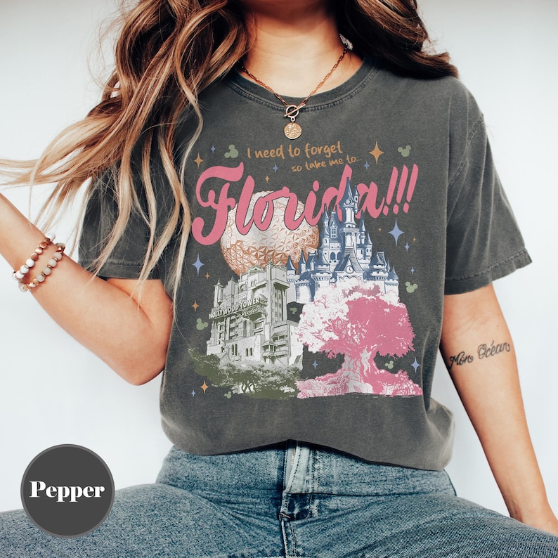 May include: Dark gray t-shirt with a colorful graphic of Florida landmarks, including a sphere, castle, and tree. The text "I need to forget so take me to... Florida!!!" is printed in pink and white.