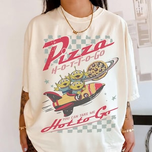 May include: Off-white t-shirt with a cartoon graphic. The design shows three green aliens in a rocket, a pizza planet, and the words "Pizza H-O-T-T-O-Go" and "You can take me Hotto Go" in red. The style is retro.