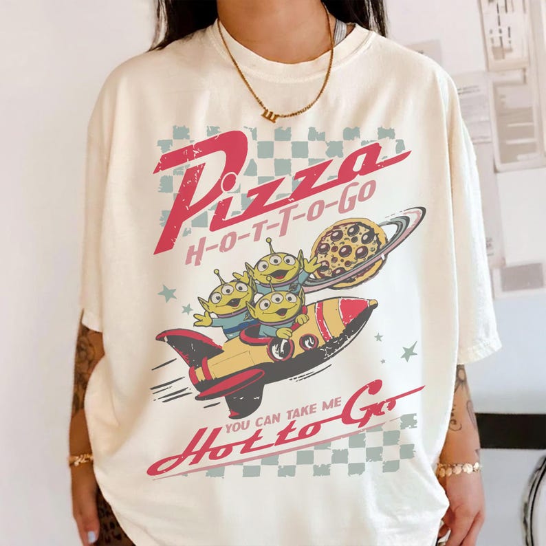 May include: Off-white t-shirt with a cartoon graphic. The design shows three green aliens in a rocket, a pizza planet, and the words "Pizza H-O-T-T-O-Go" and "You can take me Hotto Go" in red. The style is retro.
