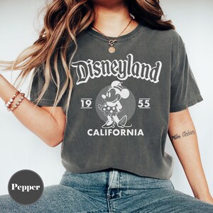 May include: Dark gray t-shirt with the word "Disneyland" in a stylized font. The shirt features a graphic of Minnie Mouse and the text "1955 CALIFORNIA". The shirt is a vintage style.