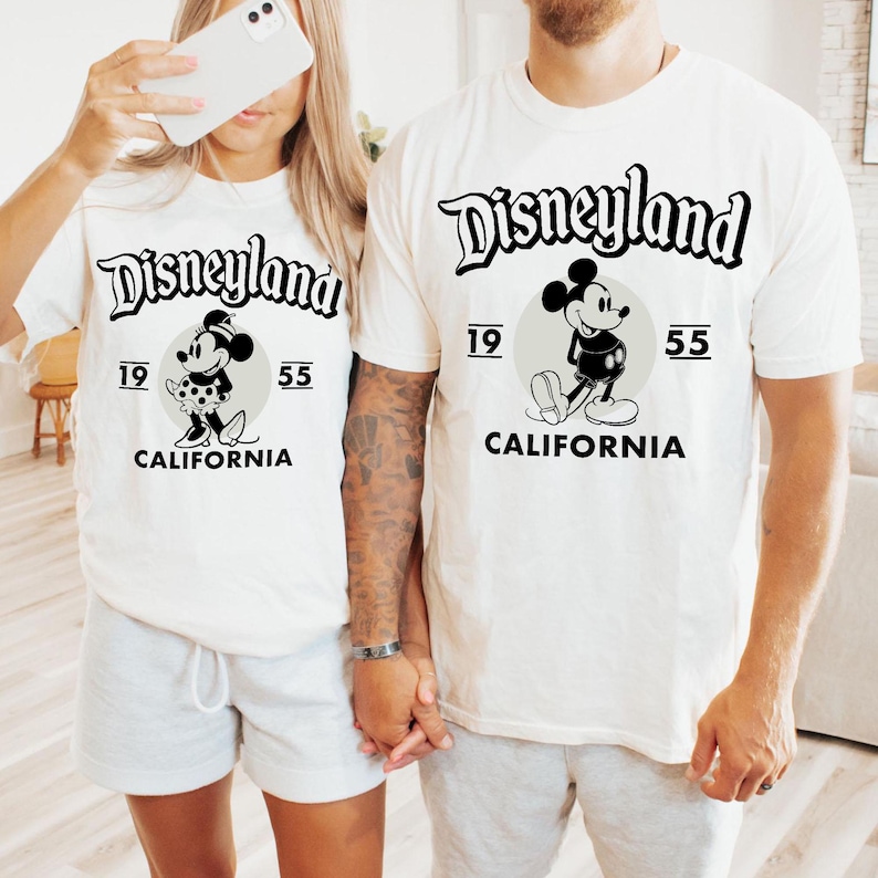 May include: Off-white t-shirts featuring the Disneyland logo in black script, with a Mickey Mouse graphic. The shirts also display the numbers "19" and "55" and the word "CALIFORNIA". The shirts are a matching set.
