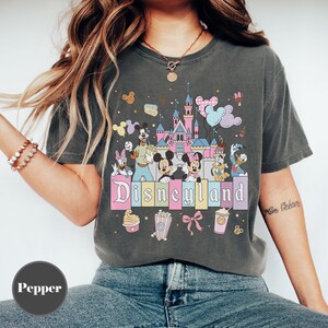 May include: Dark gray t-shirt featuring a colorful Disneyland graphic. The design includes the Disneyland castle, Mickey and Minnie Mouse, Donald Duck, and other characters. The word "Disneyland" is written in a pastel font.