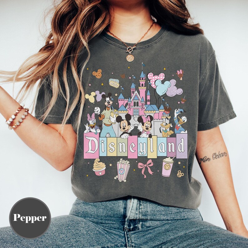 May include: Dark gray t-shirt featuring a colorful Disneyland graphic. The design includes the Disneyland castle, Mickey and Minnie Mouse, Donald Duck, and other characters. The word "Disneyland" is written in a pastel font.