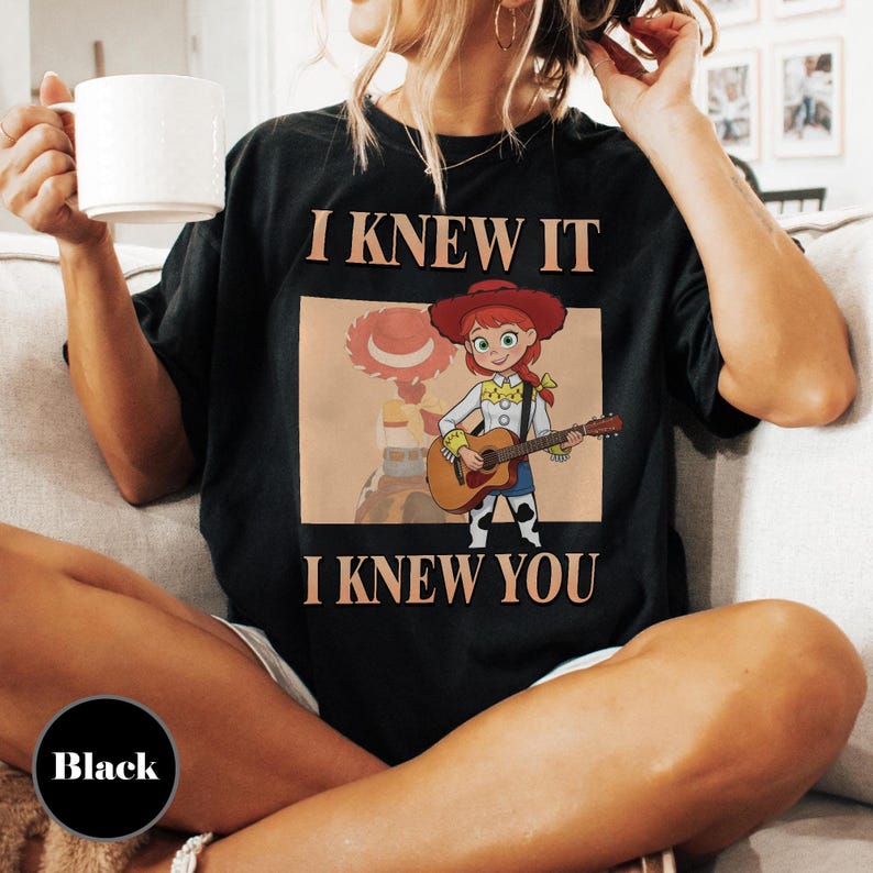 May include: A black t-shirt featuring a graphic of a cartoon cowgirl with red hair and a red hat playing an acoustic guitar. The text I KNEW IT is printed above the graphic, and the text I KNEW YOU is printed below the graphic in a tan font.