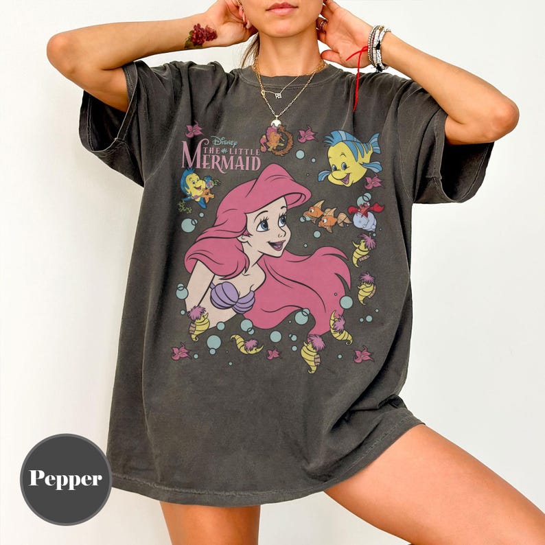 Puede incluir: Camiseta gris oscuro con Ariel de La Sirenita de Disney. El gr&aacute;fico incluye a Ariel con cabello rosa, Flounder y otras criaturas marinas. El texto "Disney The Little Mermaid" est&aacute; encima de Ariel.