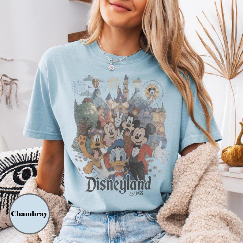 May include: Light blue t-shirt featuring a colorful graphic of Disneyland characters and landmarks. The design includes Mickey Mouse, Donald Duck, and Goofy, with the text "Disneyland Est. 1955". The shirt is a casual wear item.