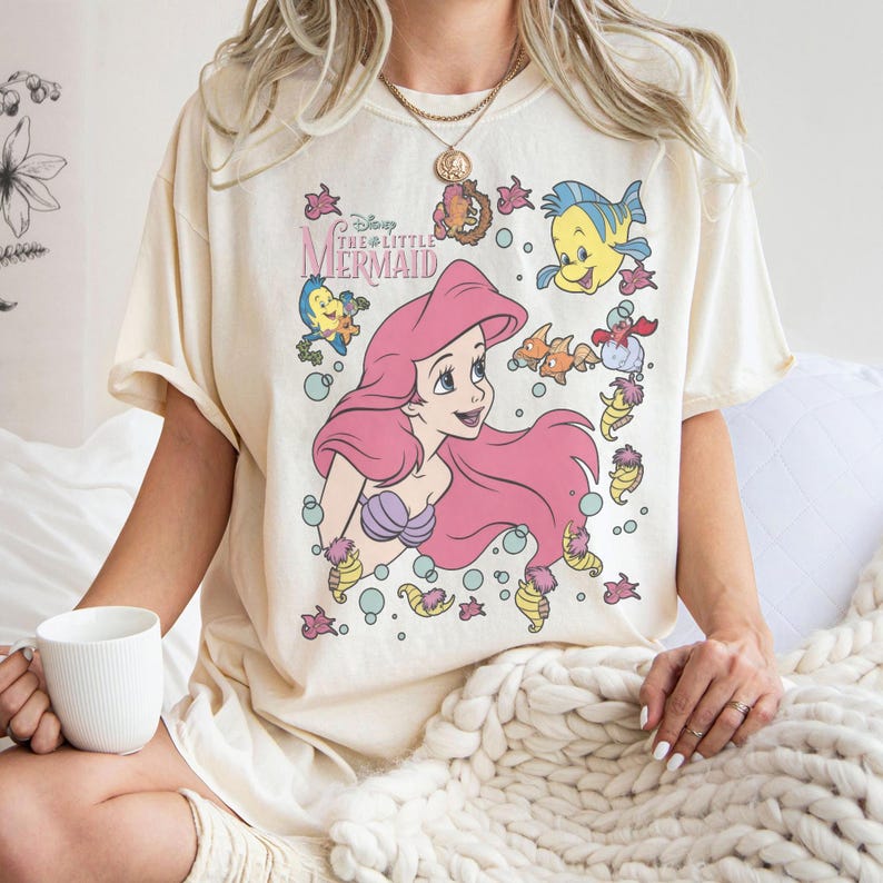 Puede incluir: Camiseta color crema con un gr&aacute;fico de Ariel de La Sirenita de Disney. El dise&ntilde;o incluye a Ariel con cabello rosa, un sujetador de concha p&uacute;rpura y varias criaturas marinas. Tambi&eacute;n es visible el texto "Disney The Little Mermaid".
