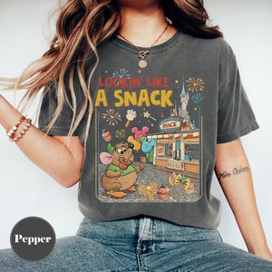 May include: Dark gray t-shirt with a vintage-style graphic. The graphic features a cartoon mouse holding balloons in front of a snack stand with the text "Lookin' Like A Snack". The design includes fireworks and a castle.