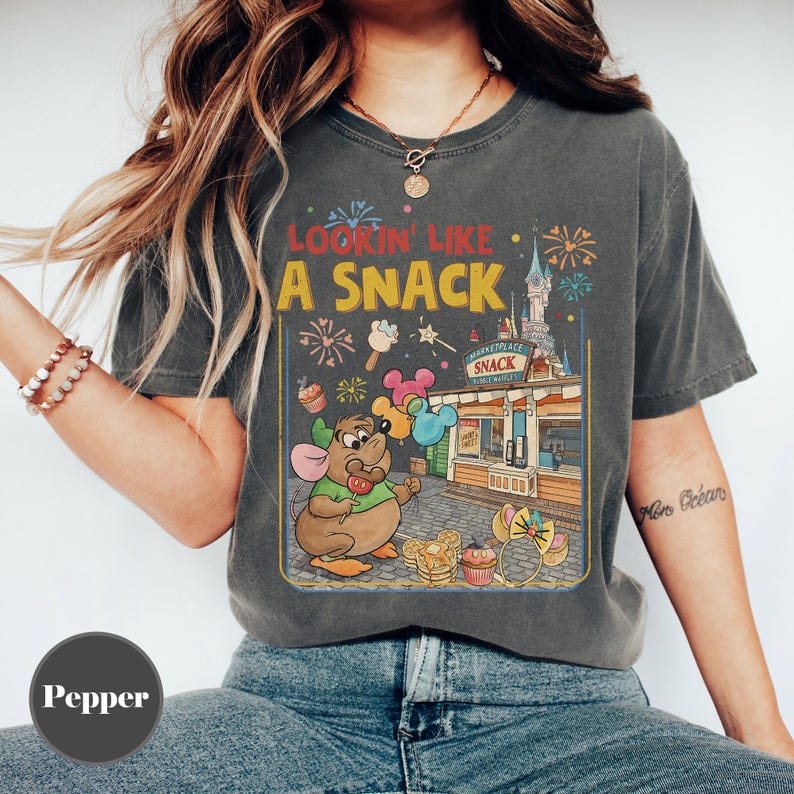 May include: Dark gray t-shirt with a vintage-style graphic. The graphic features a cartoon mouse holding balloons in front of a snack stand with the text "Lookin' Like A Snack". The design includes fireworks and a castle.