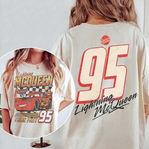 May include: Cream-colored t-shirt featuring a graphic of Lightning McQueen from the Cars movie. The front design includes the character, the number 95, and the words "Lightning McQueen" and "Think Fast." The back displays a large number 95.