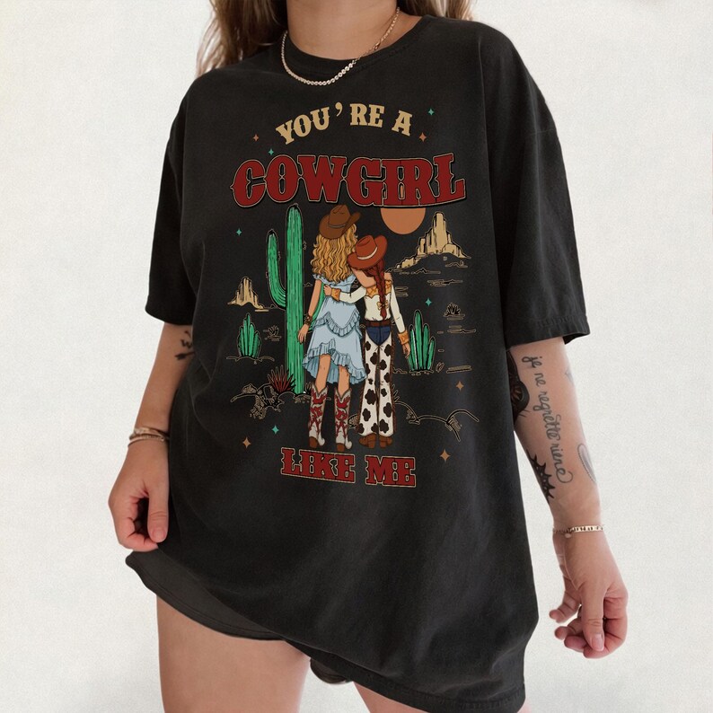 May include: A black short-sleeve t-shirt featuring a graphic print of two figures in cowboy hats and boots standing in a desert landscape with cacti and mountains. The text "YOU'RE A COWGIRL LIKE ME" is printed in red above and below the illustration.