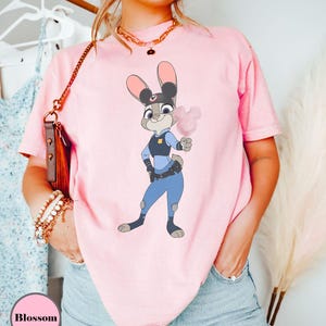 Puede incluir: Una camiseta rosa claro con una ilustraci&oacute;n de dibujos animados de Judy Hopps de Zootopia, sosteniendo algod&oacute;n de az&uacute;car rosa. El personaje lleva un uniforme de polic&iacute;a. La palabra "Blossom" est&aacute; en un c&iacute;rculo rosa en la parte inferior izquierda.