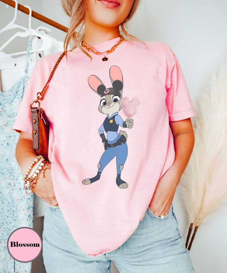Puede incluir: Una camiseta rosa claro con una ilustraci&oacute;n de dibujos animados de Judy Hopps de Zootopia, sosteniendo algod&oacute;n de az&uacute;car rosa. El personaje lleva un uniforme de polic&iacute;a. La palabra "Blossom" est&aacute; en un c&iacute;rculo rosa en la parte inferior izquierda.