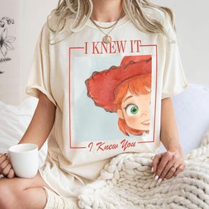 K&ouml;nnte beinhalten: Ein cremefarbenes Kurzarm-T-Shirt mit einem Grafikdruck einer Zeichentrickfigur mit roten Haaren und einem roten Cowboyhut. Der Text "I KNEW IT" ist &uuml;ber der Figur und "I Knew You" darunter gedruckt, alles innerhalb eines roten Rahmens.