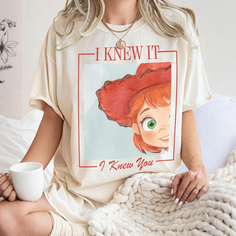 K&ouml;nnte beinhalten: Ein cremefarbenes Kurzarm-T-Shirt mit einem Grafikdruck einer Zeichentrickfigur mit roten Haaren und einem roten Cowboyhut. Der Text "I KNEW IT" ist &uuml;ber der Figur und "I Knew You" darunter gedruckt, alles innerhalb eines roten Rahmens.