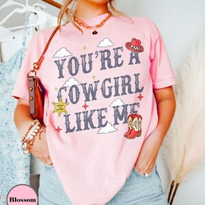Puede incluir: Una camiseta de manga corta de color rosa claro con el texto "YOU'RE A COWGIRL LIKE ME" en letras gris oscuro. El dise&ntilde;o incluye ilustraciones de un sombrero de vaquero rojo, una estrella de sheriff amarilla, botas rojas y nubes blancas.