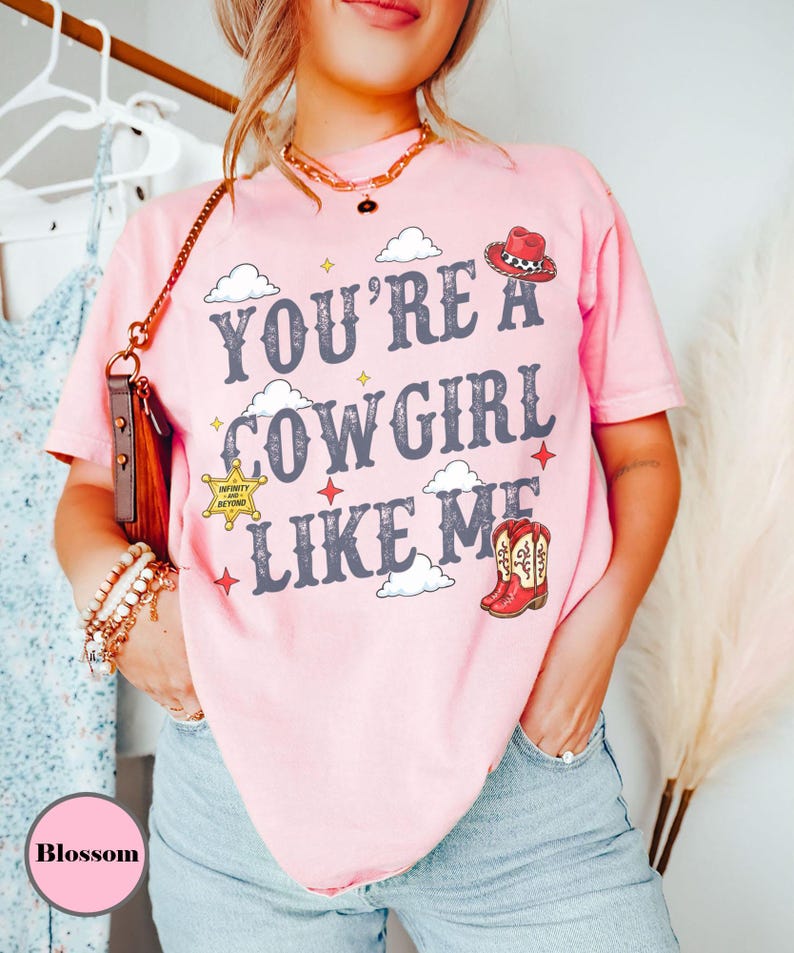 Puede incluir: Una camiseta de manga corta de color rosa claro con el texto "YOU'RE A COWGIRL LIKE ME" en letras gris oscuro. El dise&ntilde;o incluye ilustraciones de un sombrero de vaquero rojo, una estrella de sheriff amarilla, botas rojas y nubes blancas.