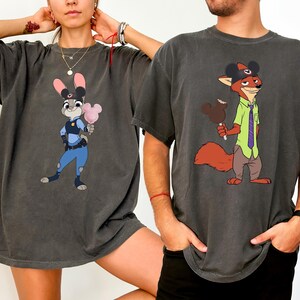 Puede incluir: Dos camisetas gris carb&oacute;n con personajes de dibujos animados. Una camiseta muestra a Judy Hopps sosteniendo algod&oacute;n de az&uacute;car, la otra a Nick Wilde con un helado de Mickey Mouse. Ambas camisetas tienen un aspecto vintage.