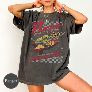 May include: Dark grey t-shirt with a graphic featuring the text "Pizza H-O-T-T-O-Go" in red and white. The design includes cartoon aliens in a rocket ship with a pizza planet. The shirt has a vintage, distressed appearance.