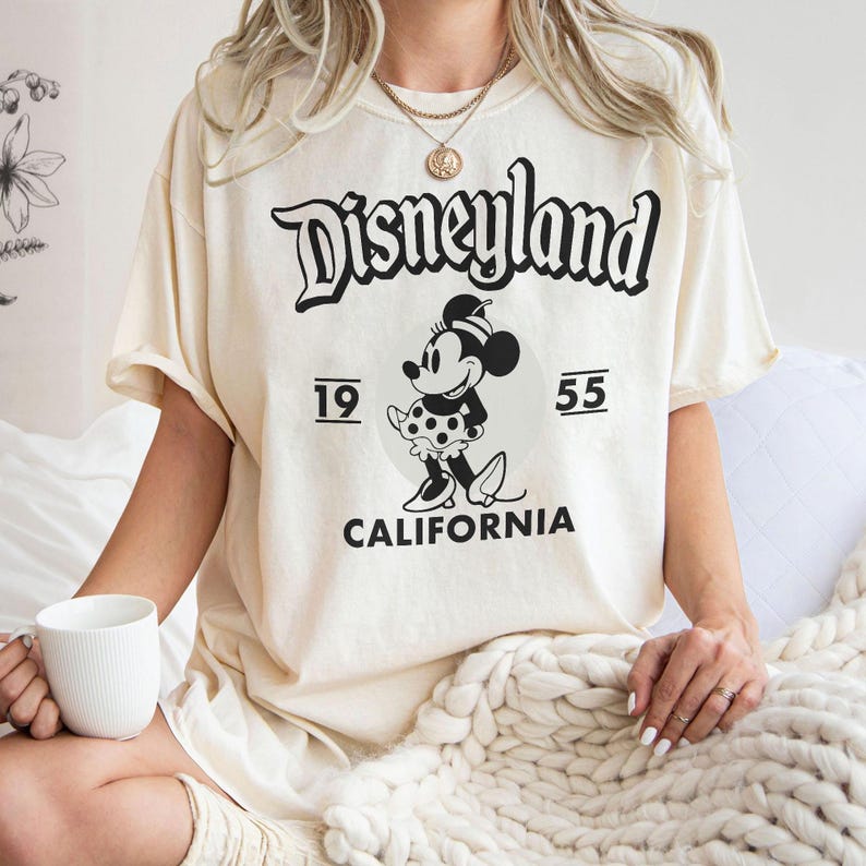 May include: Cream-colored t-shirt featuring a black graphic of the Disneyland logo, Minnie Mouse, and the text "1955 CALIFORNIA." The shirt has short sleeves and a relaxed fit. A white mug is held in the left hand.