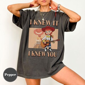 May include: A charcoal gray short-sleeve t-shirt featuring a graphic of a cowgirl character playing an acoustic guitar. The text "I KNEW IT" appears above the graphic, and "I KNEW YOU" appears below it in a bold, tan-colored serif font.