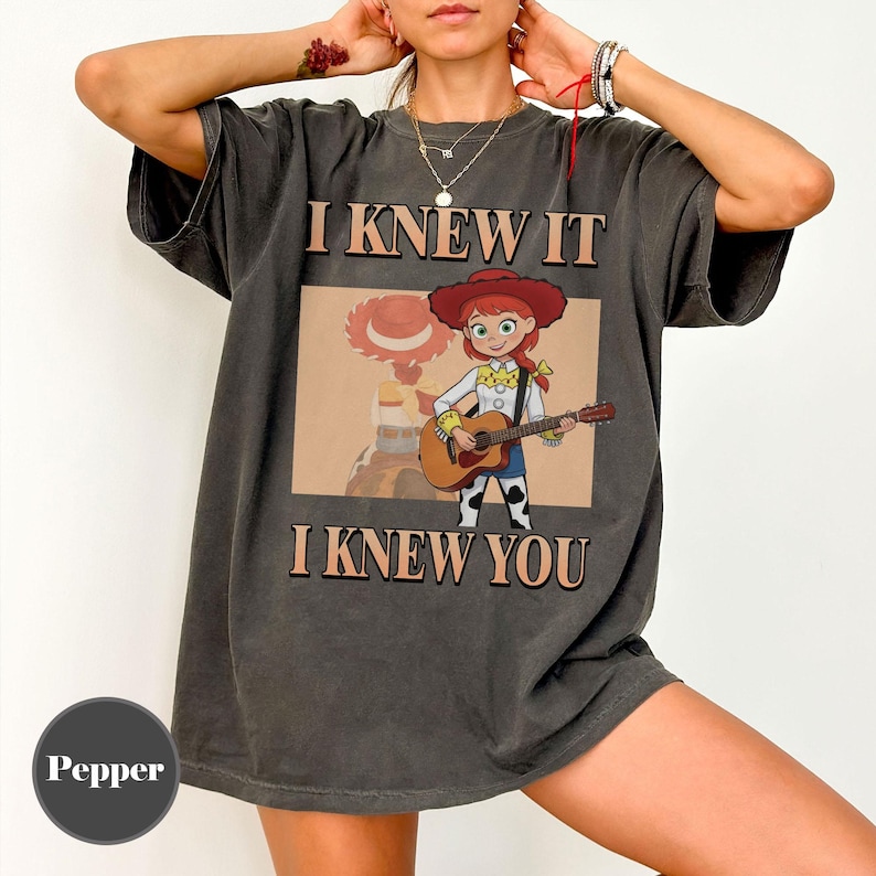 May include: A charcoal gray short-sleeve t-shirt featuring a graphic of a cowgirl character playing an acoustic guitar. The text "I KNEW IT" appears above the graphic, and "I KNEW YOU" appears below it in a bold, tan-colored serif font.