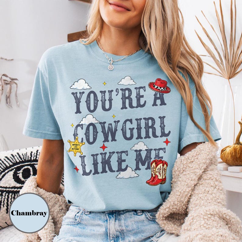 Puede incluir: Una camiseta de manga corta azul claro con el texto "YOU'RE A COWGIRL LIKE ME" en letras azul oscuro. El dise&ntilde;o incluye ilustraciones de un sombrero de vaquero rojo, una estrella de sheriff amarilla, una bota roja y nubes blancas.
